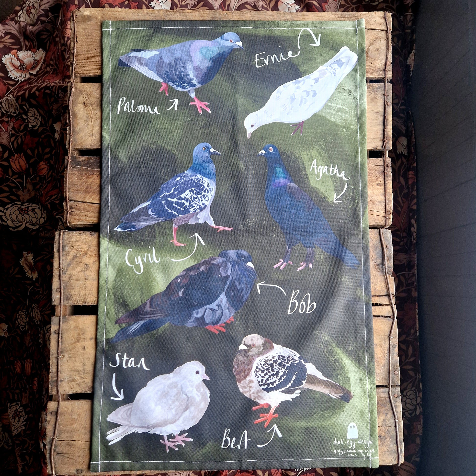 A green tea towel with seven different pigeons on lies on a wooden plank background. 