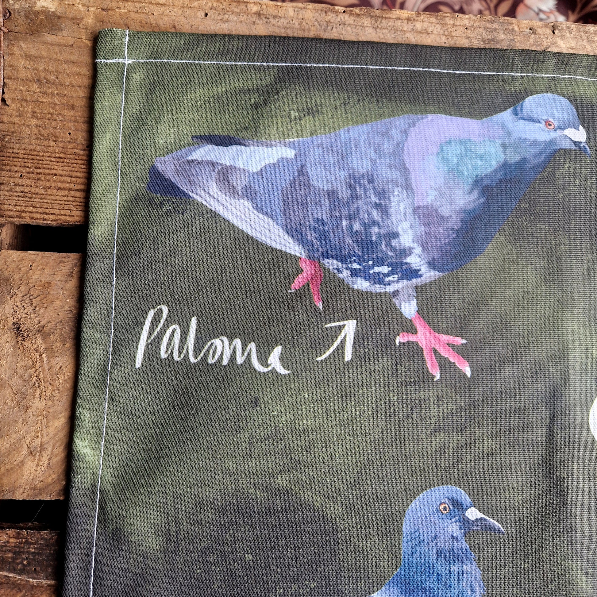 A close look at the top left of the tea towel showing the detail in the pigeon illustrations. Behind the tea towel you can see a wooden planks background.
