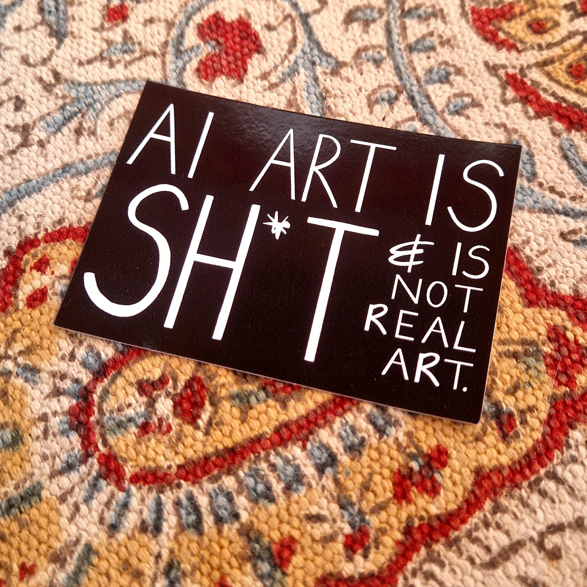 A black rectangular sticker featuring the words 'AI ART IS SHIT & IS NOT REAL ART. ' in white handwriting. The sticker sits on a patterned fabric background.