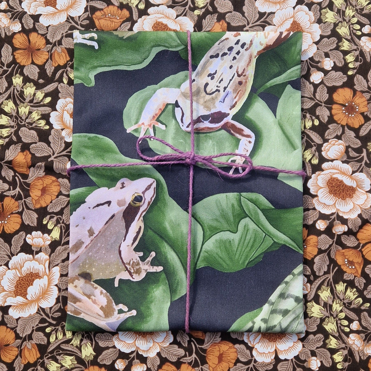A folded blue and green teatowel with two frogs visible on it sits wrapped in a purple twine on top of a floral patterned background.