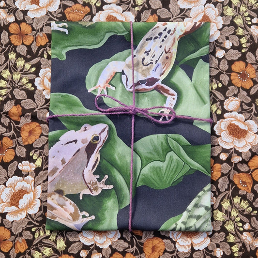 A folded blue and green teatowel with two frogs visible on it sits wrapped in a purple twine on top of a floral patterned background.