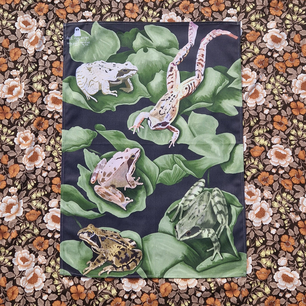 A deep blue teatowel sits on a floral patterned background. The tea towel is covered in lily pads with five different frogs hanging out on it.