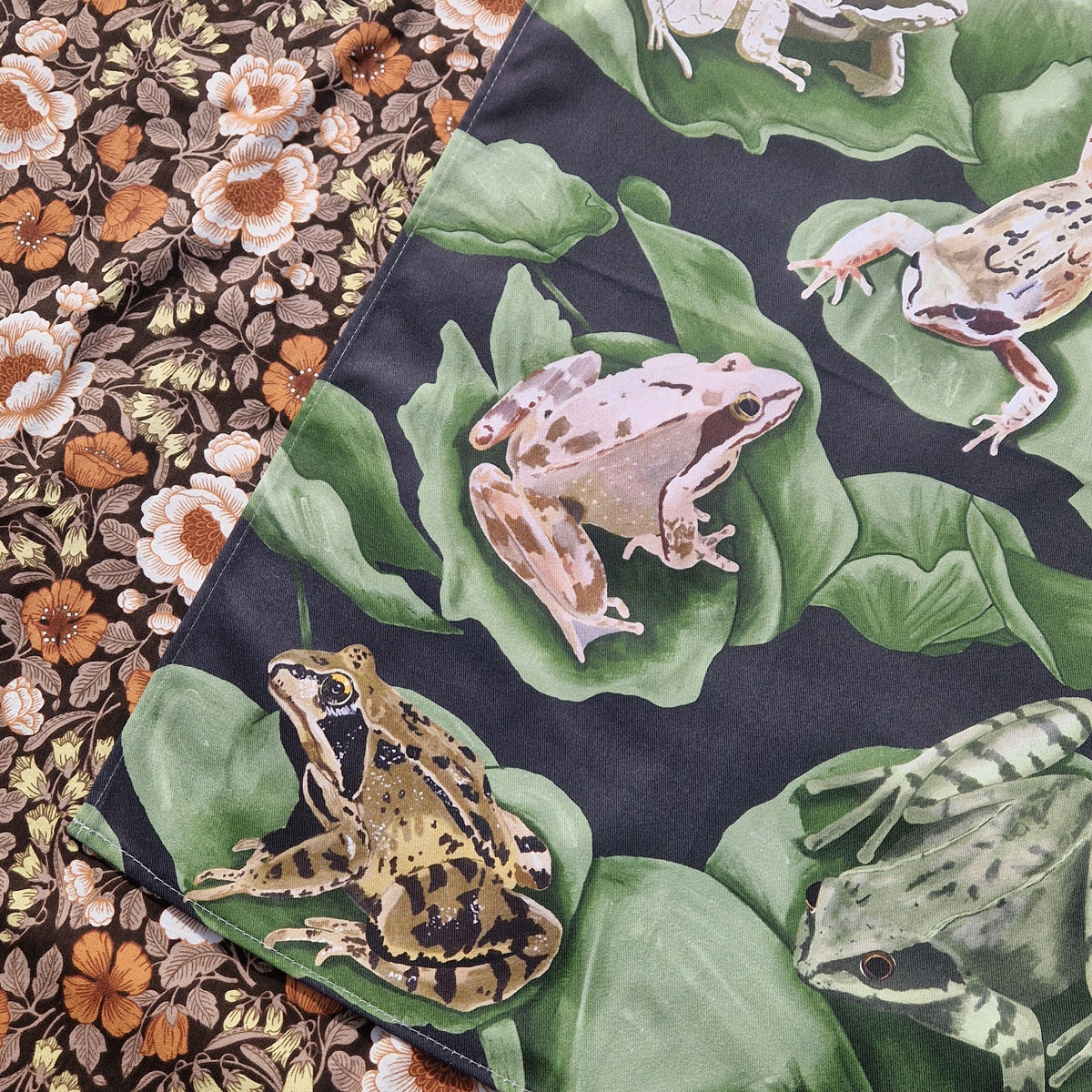 A close up of the bottom left of the teatowel showing the detail in the frog illustrations. To the left of the deep blue teatowel is a floral patterned background.