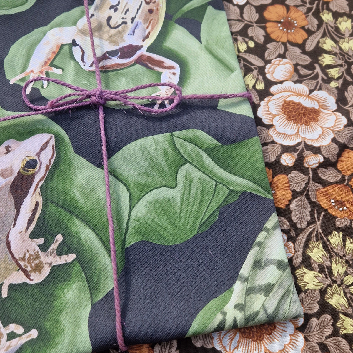 A close up of the folded dark blue teatowel showing the detail in the frog and lilypad design. To the right of the tea towel is a floral patterned fabric.