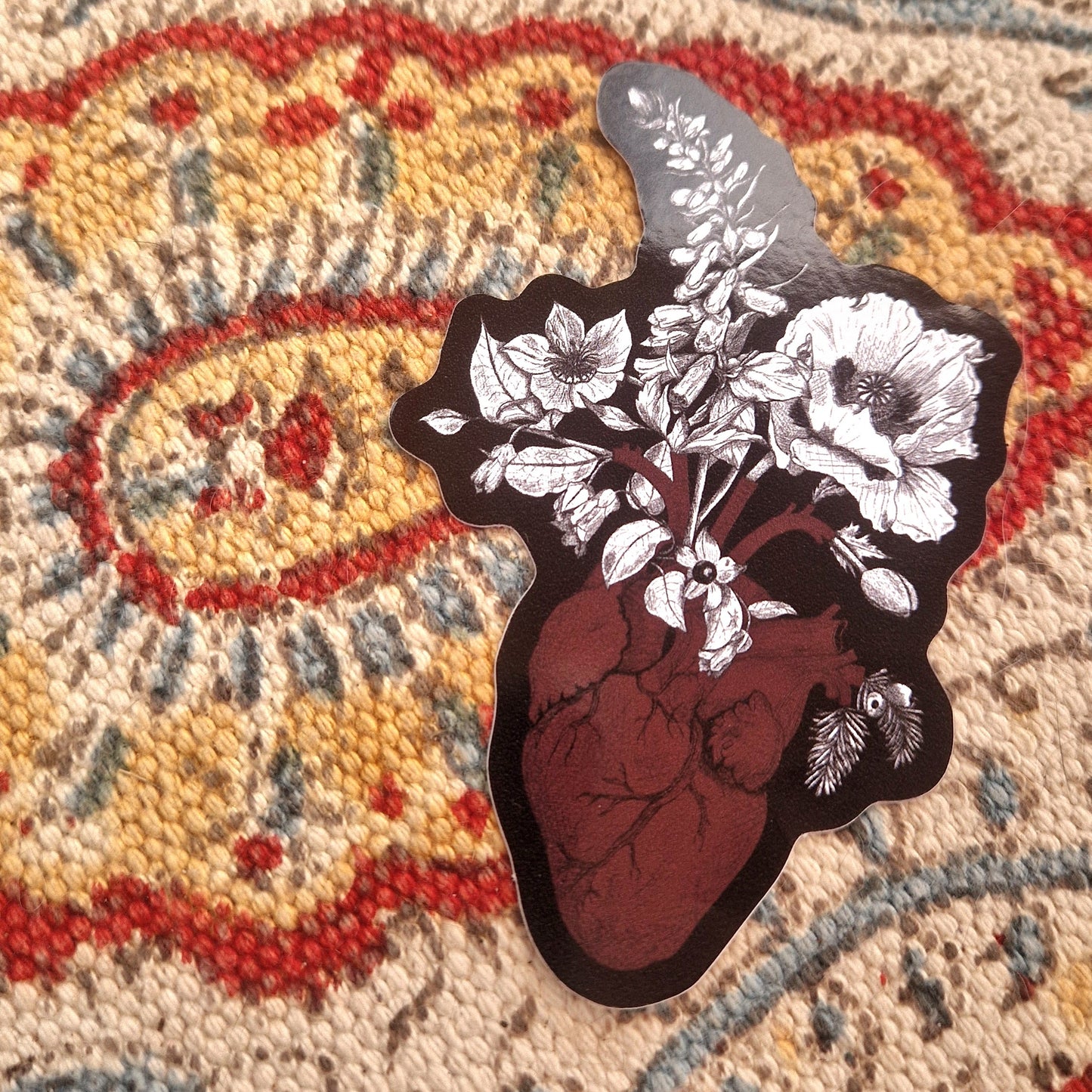 A sticker with a black background featuring a red anatomical heart and with white and black poisonous plants coming out of it sits on a patterned fabric background. 