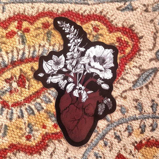 A sticker with a black background featuring a red anatomical heart and with white and black poisonous plants coming out of it sits on a patterned fabric background. 
