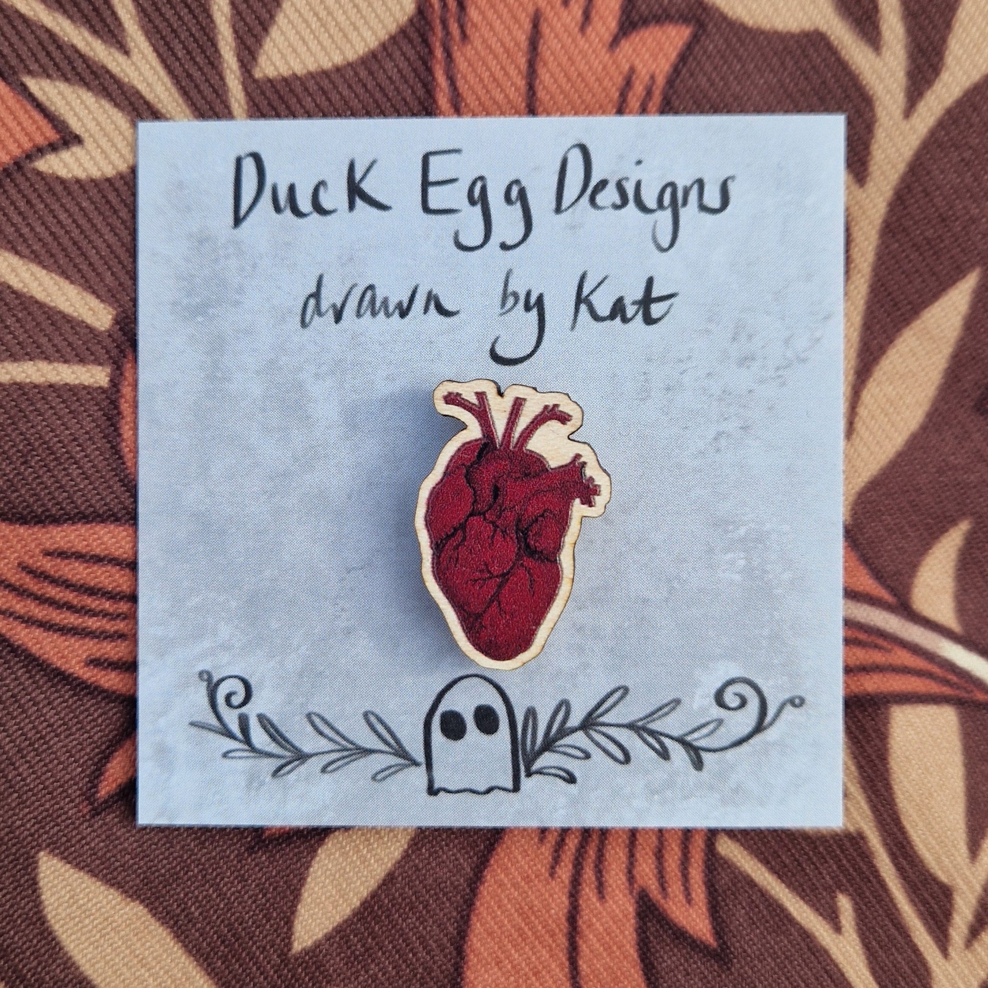 A rich red anatomical heart pin badge sits on a grey backing card with the words 'Duck Egg Designs drawn by Kat' in black across the top. Behind the backing card is a brown patterned fabric.