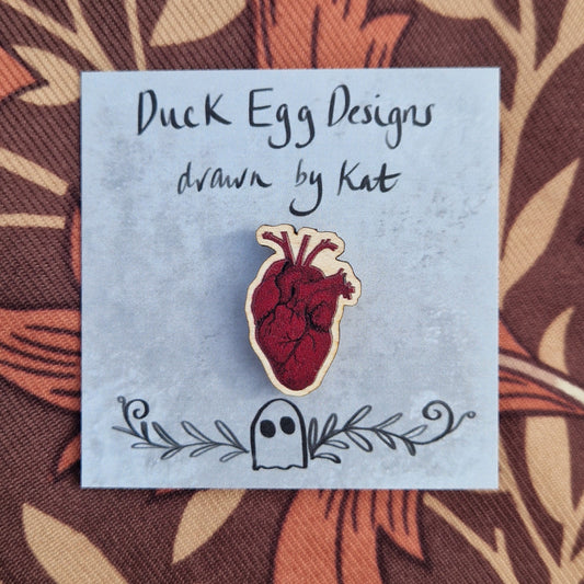 A rich red anatomical heart pin badge sits on a grey backing card with the words 'Duck Egg Designs drawn by Kat' in black across the top. Behind the backing card is a brown patterned fabric.