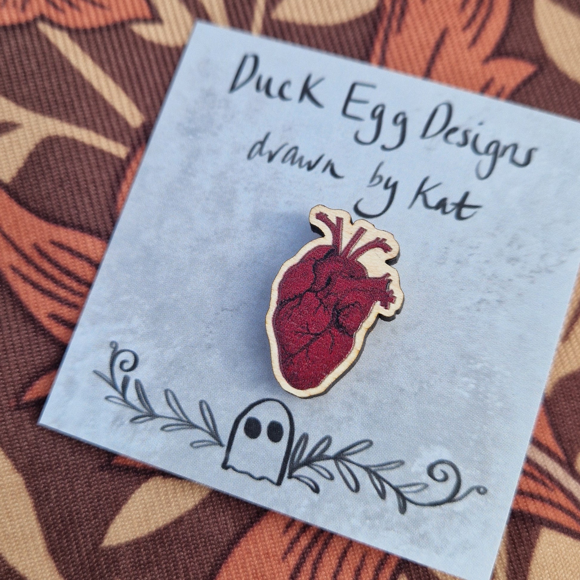 A rich red anatomical heart pin badge sits on a grey backing card with the words 'Duck Egg Designs drawn by Kat' in black across the top. Behind the backing card is a brown patterned fabric.