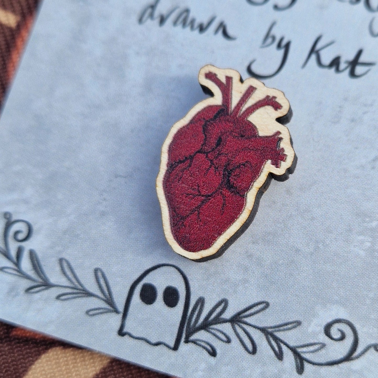 A rich red anatomical heart pin badge sits on a grey backing card with the words 'Duck Egg Designs drawn by Kat' in black across the top. Behind the backing card is a brown patterned fabric.