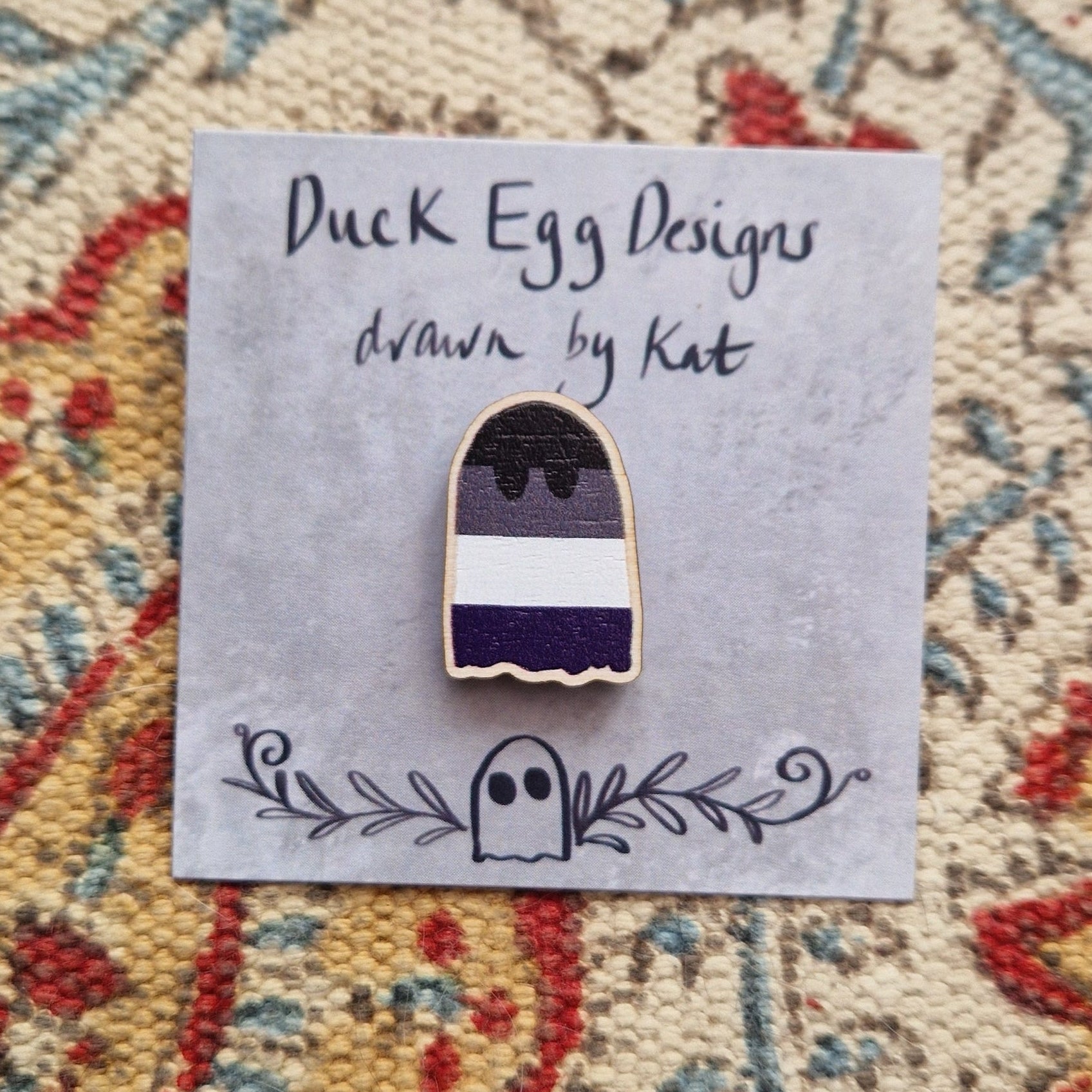 A striped ghost pin featuring the asexual flag sits on a grey backing card with the words 'Duck Egg Designs drawn by Kat' written across the top. Behind the backing card you can see a patterned fabric background.