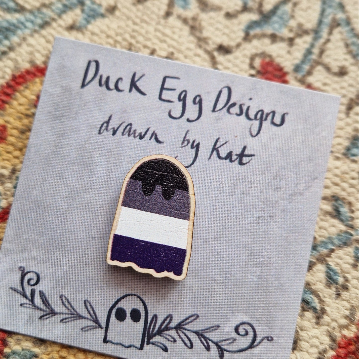A close up of the asexual flag ghost pin, showing the texture of the wood. The pin sits on a grey backing card in front of a patterned fabric background.