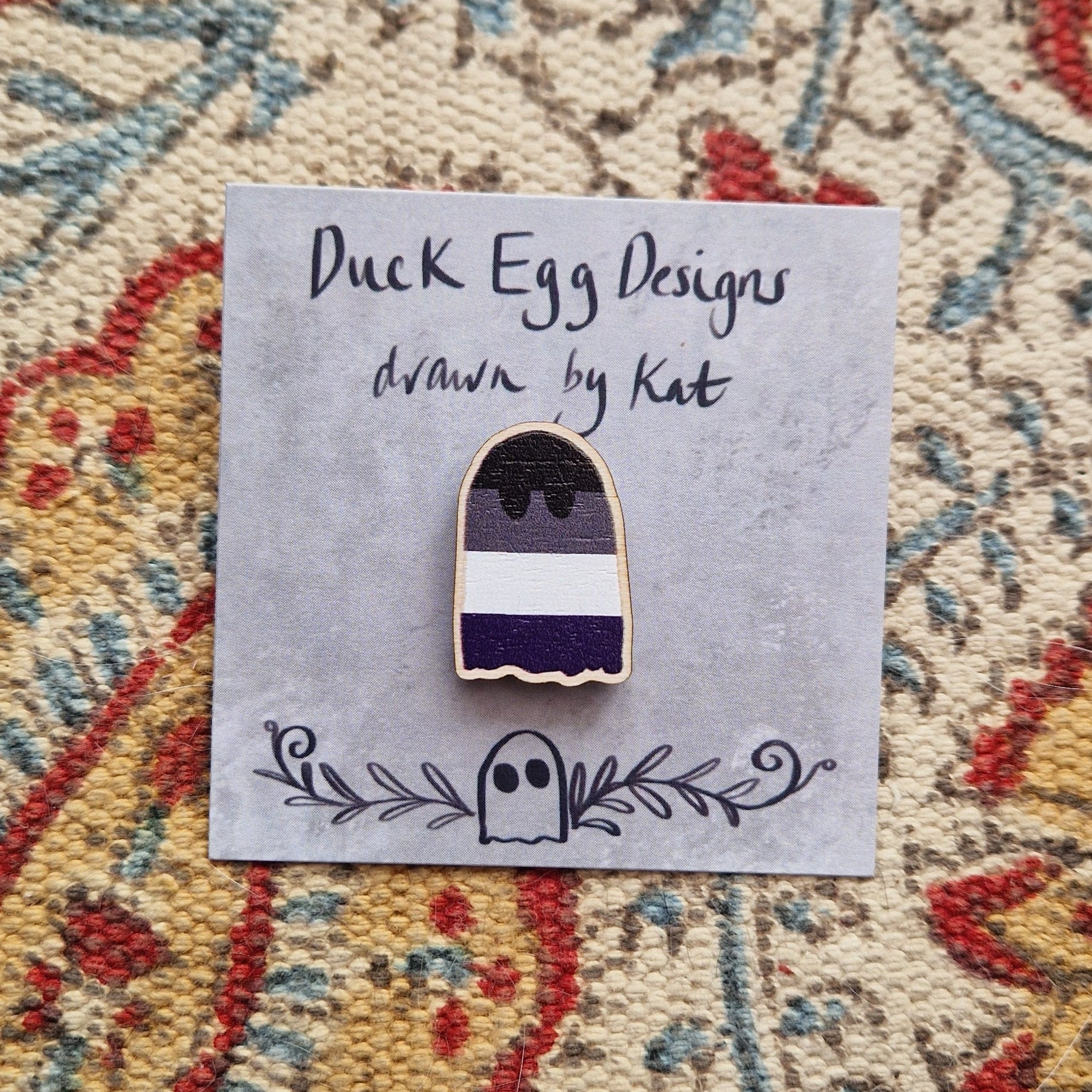 A striped ghost pin featuring the asexual flag sits on a grey backing card with the words 'Duck Egg Designs drawn by Kat' written across the top. Behind the backing card you can see a patterned fabric background.