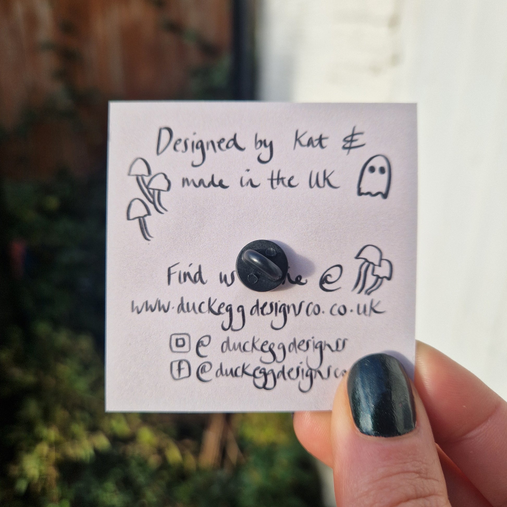 The back of the pin badge, showing the black rubber pin back on the dusky pink of the backing card. The backing card is held by a white hand wearing black nail polish and featuring the words 'Designed by Kat & made in the UK' with website and social media information below.