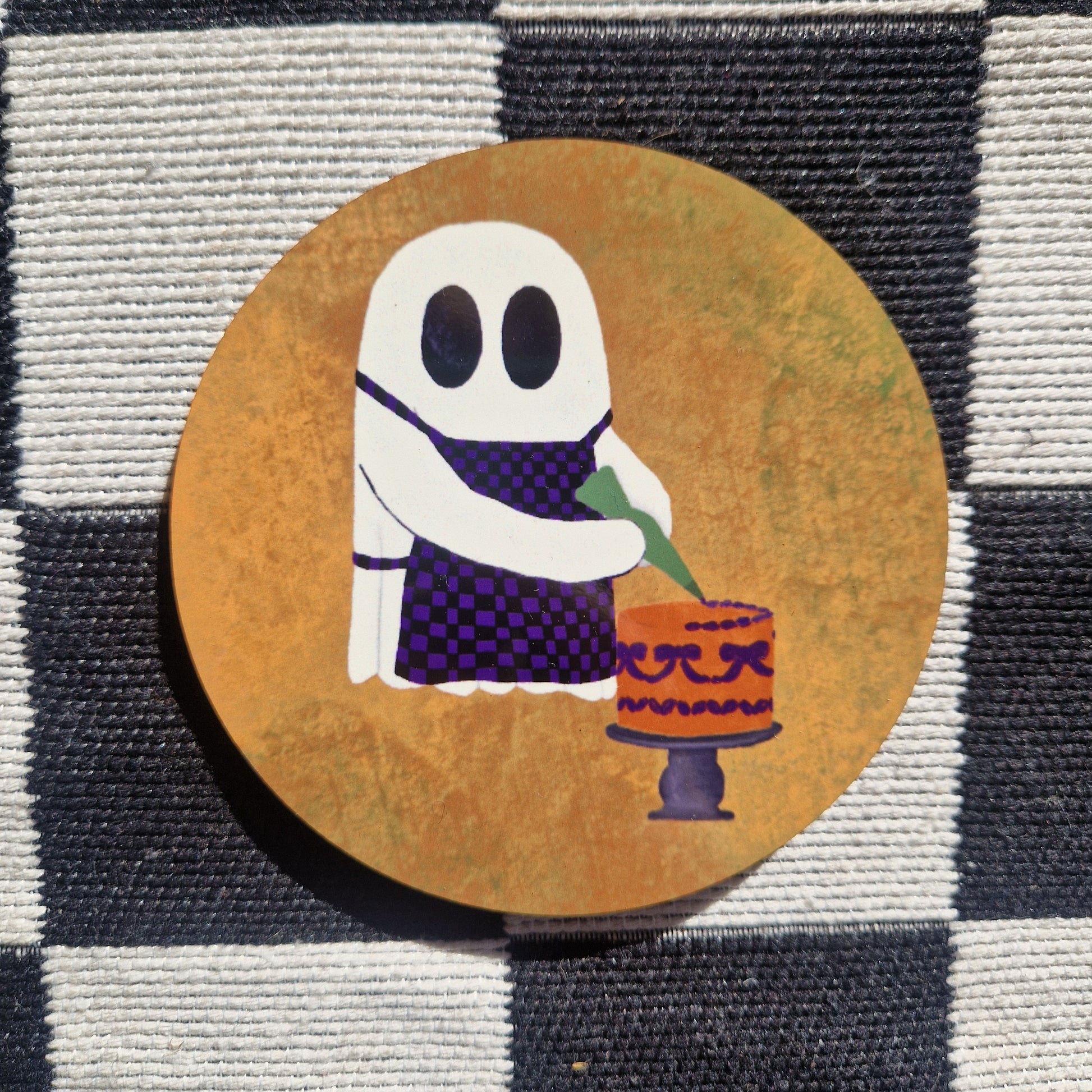 A round orange coaster featuring a ghost wearing a black and purple checkerboard apron icing an orange and purple cake. the coaster sits on a white and black checkerboard fabric.