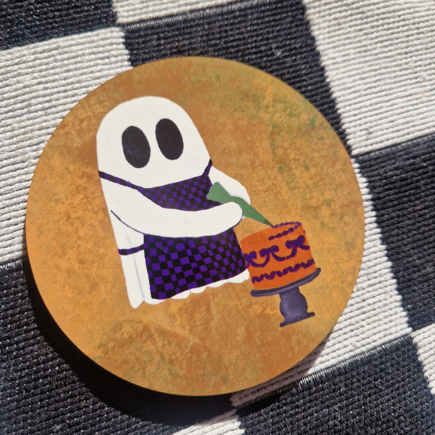 An orange round coaster sits on a black and white checkerboard fabric. The coaster features a ghost wearing a black and purple checkerboard apron, icing an orange and purple cake.