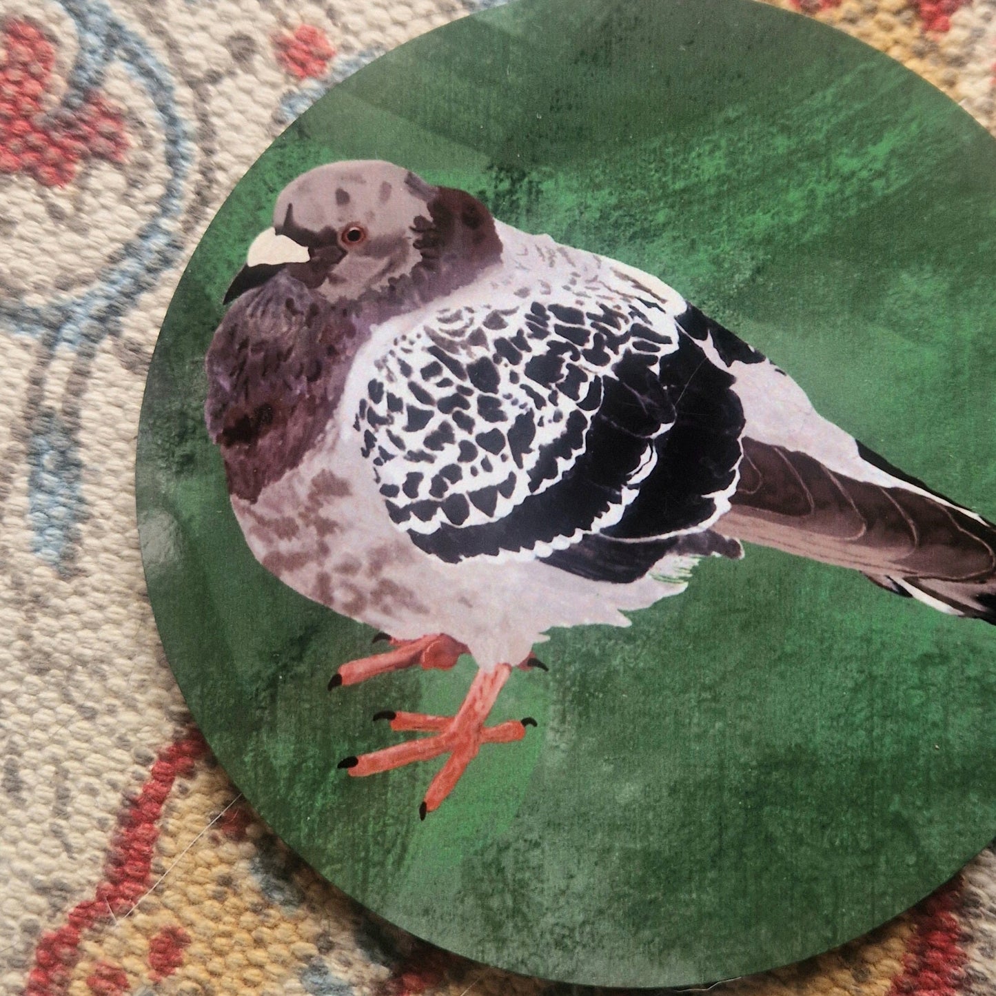 A close up of the round green coaster featuring a chunky grey pigeon illustration. The coaster sits on a patterned fabric background.