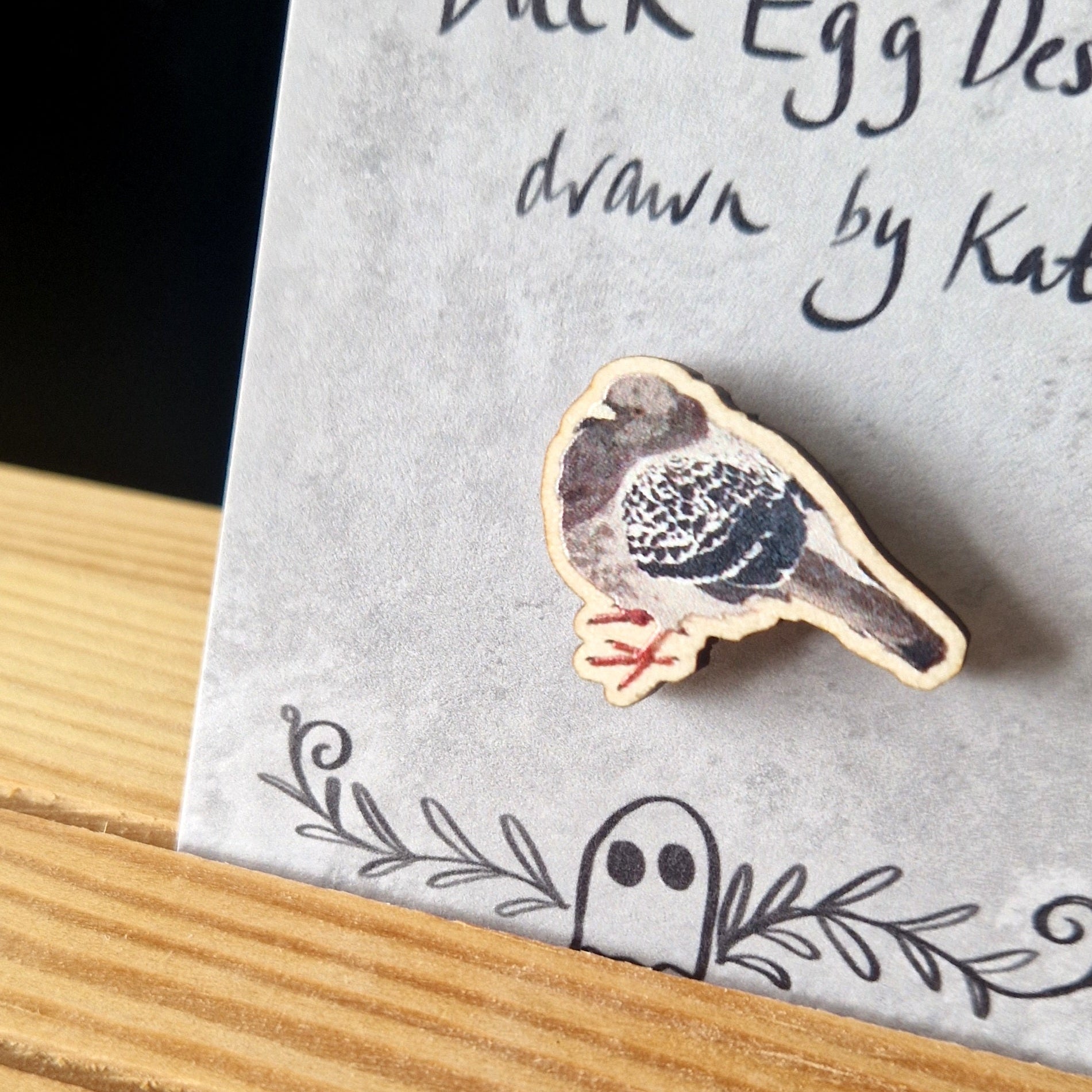 A round pigeon pin badge sits on a grey backing card in front of a black background. The pigeon is grey and black with orange feet.
