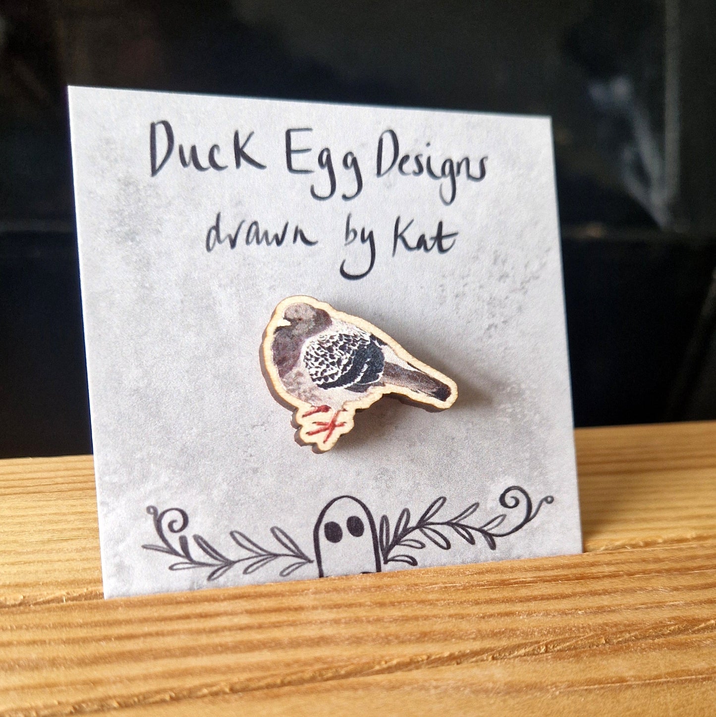 A round pigeon pin badge sits on a grey backing card in front of a black background. The pigeon is grey and black with orange feet.