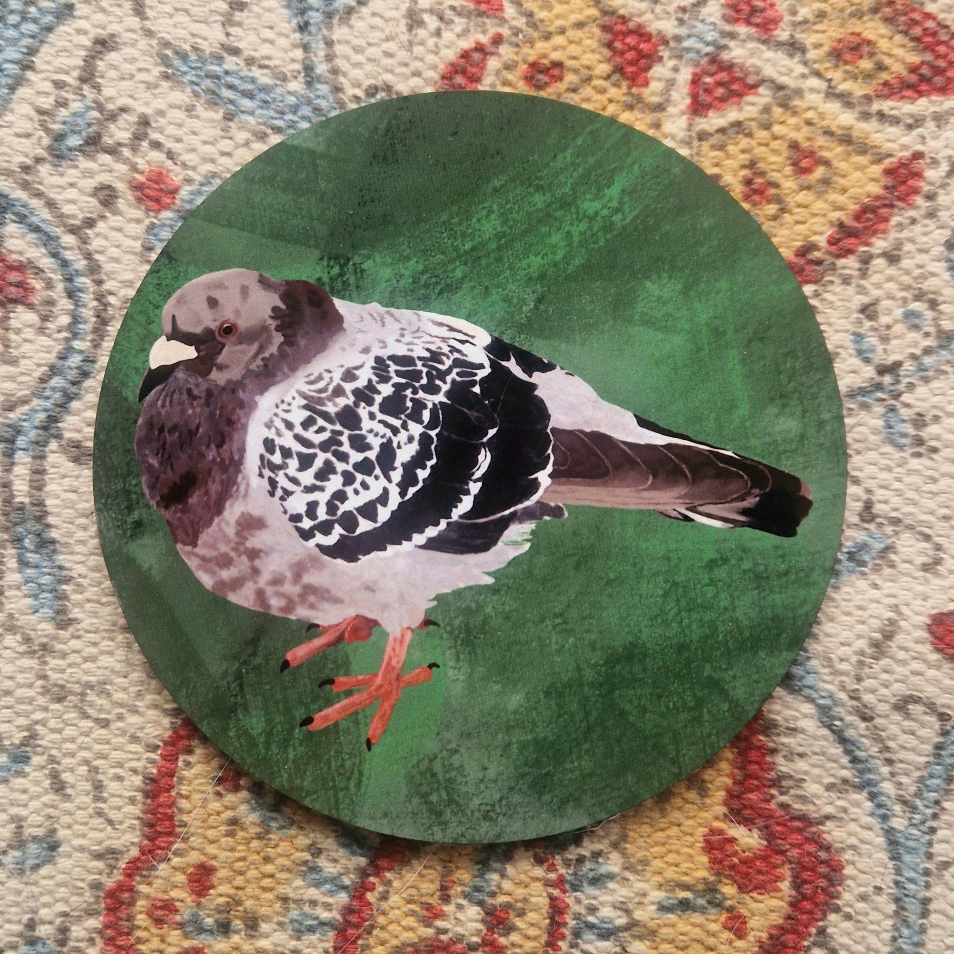 A green round coaster with a grey chunky pigeon illustration on it. The coaster sits on a cream, yellow, blue and red patterned fabric.