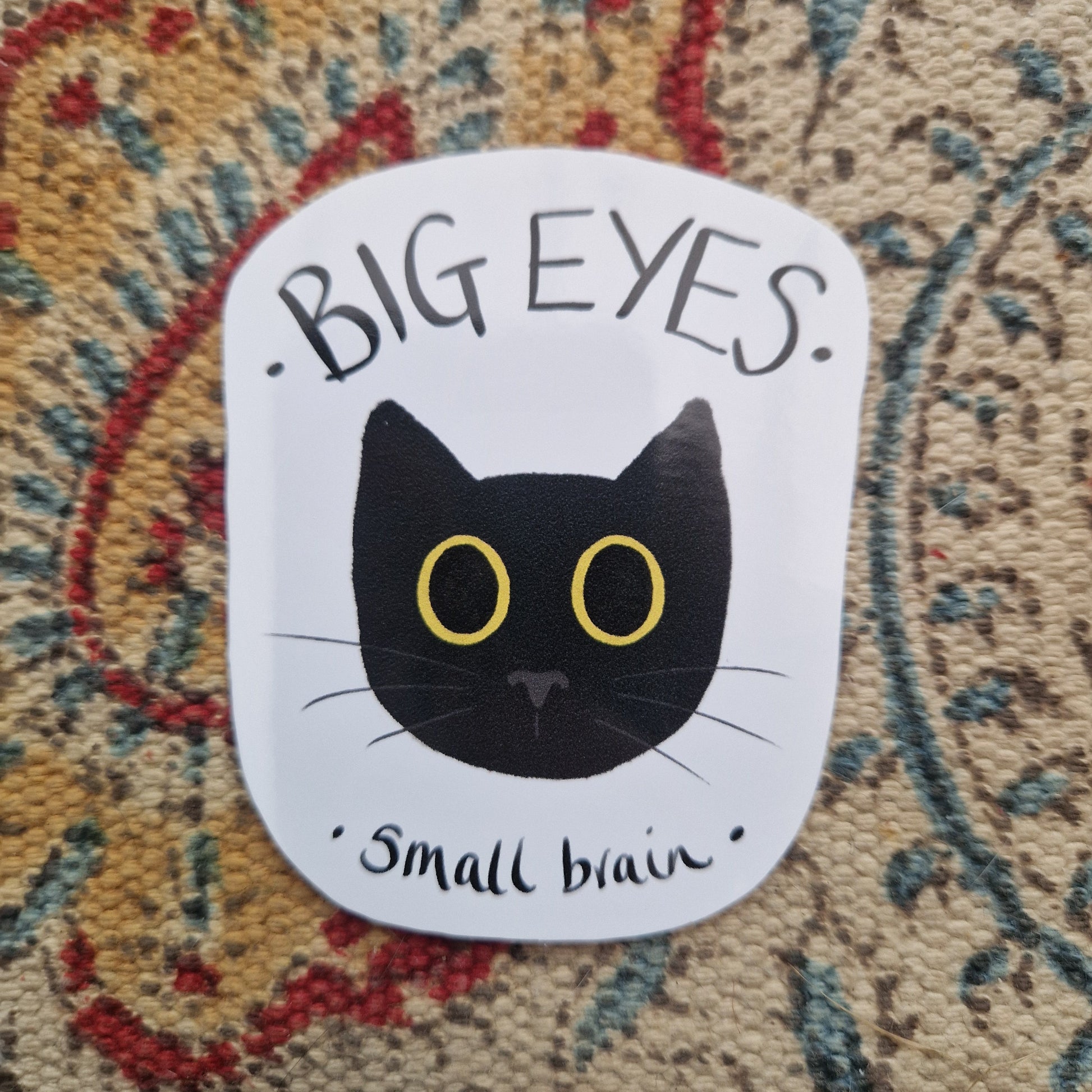 A white sticker with a big yellow eyed black cat surrounded by the words 'BIG EYES small brain' in black handwriting. The sticker sits on a patterned fabric background.