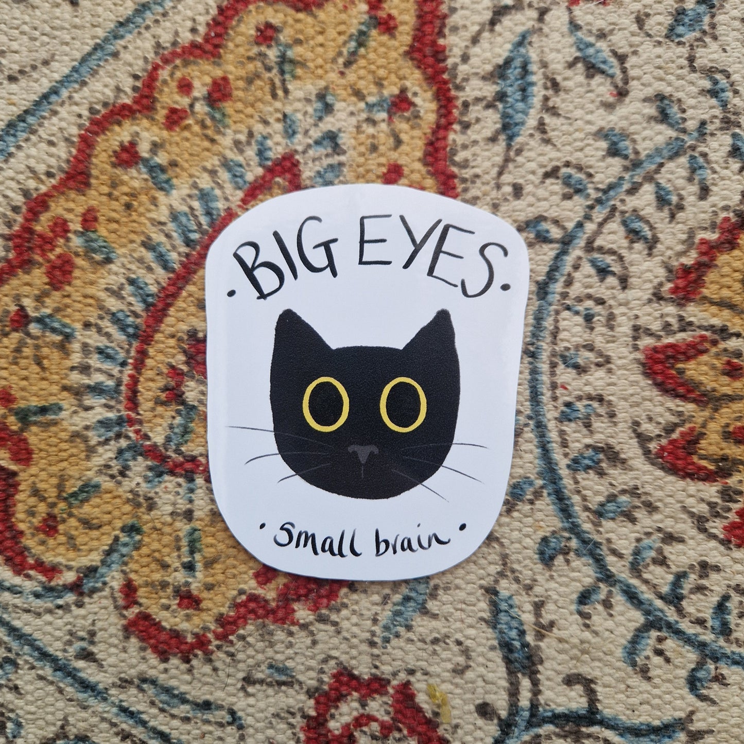 A white sticker sits on a patterned fabric background. The sticker has a black cat head with big yellow eyes in the middle with the words 'BIG EYES small brain' in black handwriting.