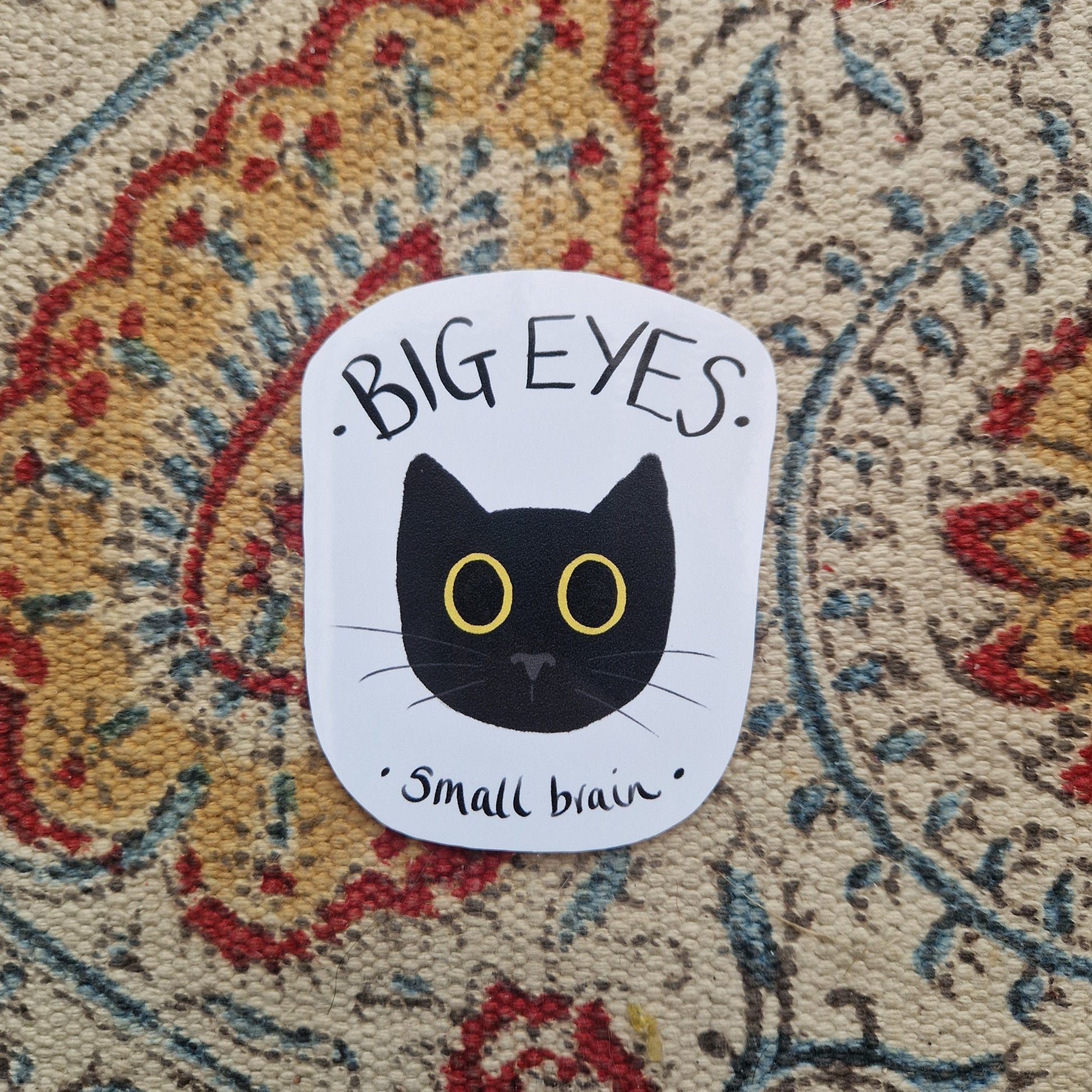 A white sticker sits on a patterned fabric background. The sticker has a black cat head with big yellow eyes in the middle with the words 'BIG EYES small brain' in black handwriting.
