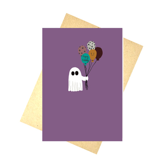 A rich purple card with a mall white ghost holding a bunch of multicoloured balloons, one of which reads 'Happy Birthday' in black handwriting. Behind the card is a brown envelope and a white background.