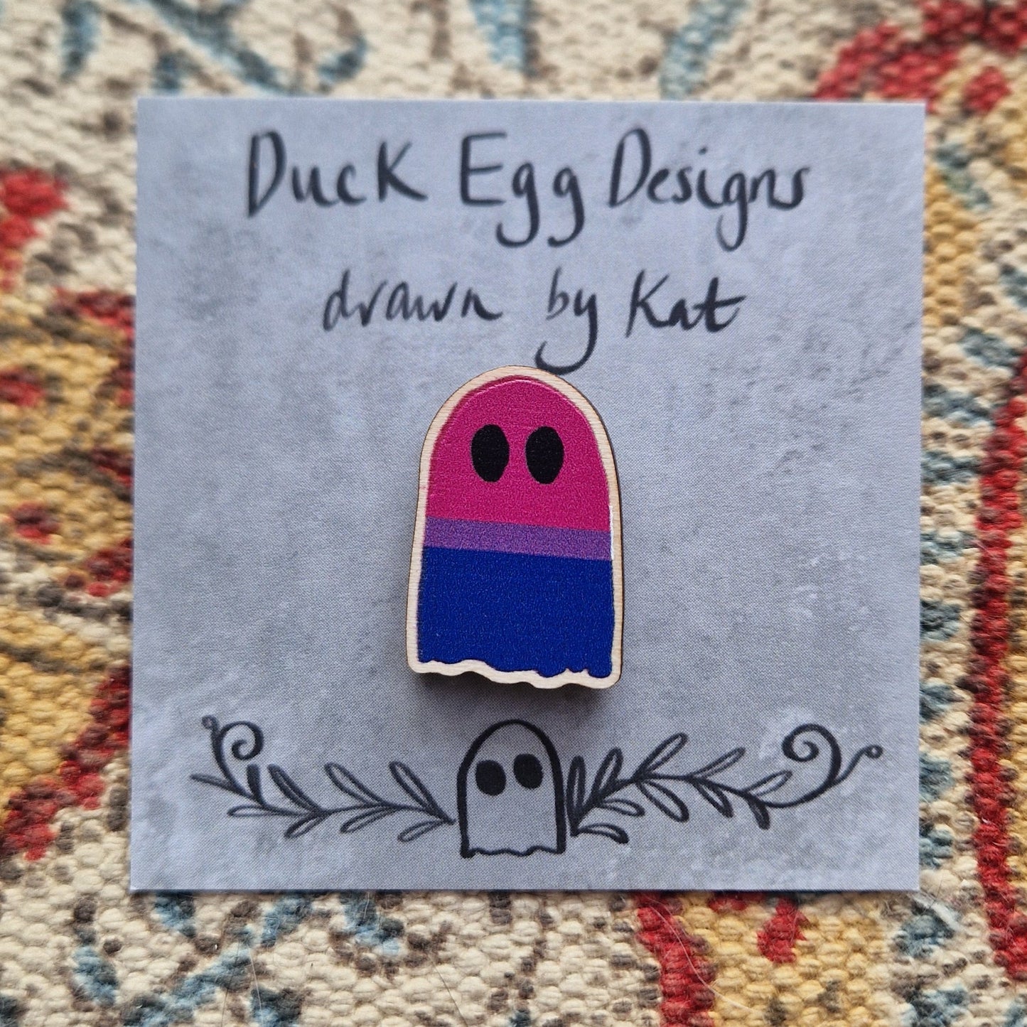 A ghost pin featuring the bisexual flag (with stripes of pink, purple and blue) sits on a grey backing card with the words 'Duck Egg Designs drawn by Kat' across the top. Behind the backing card is a patterned fabric background.