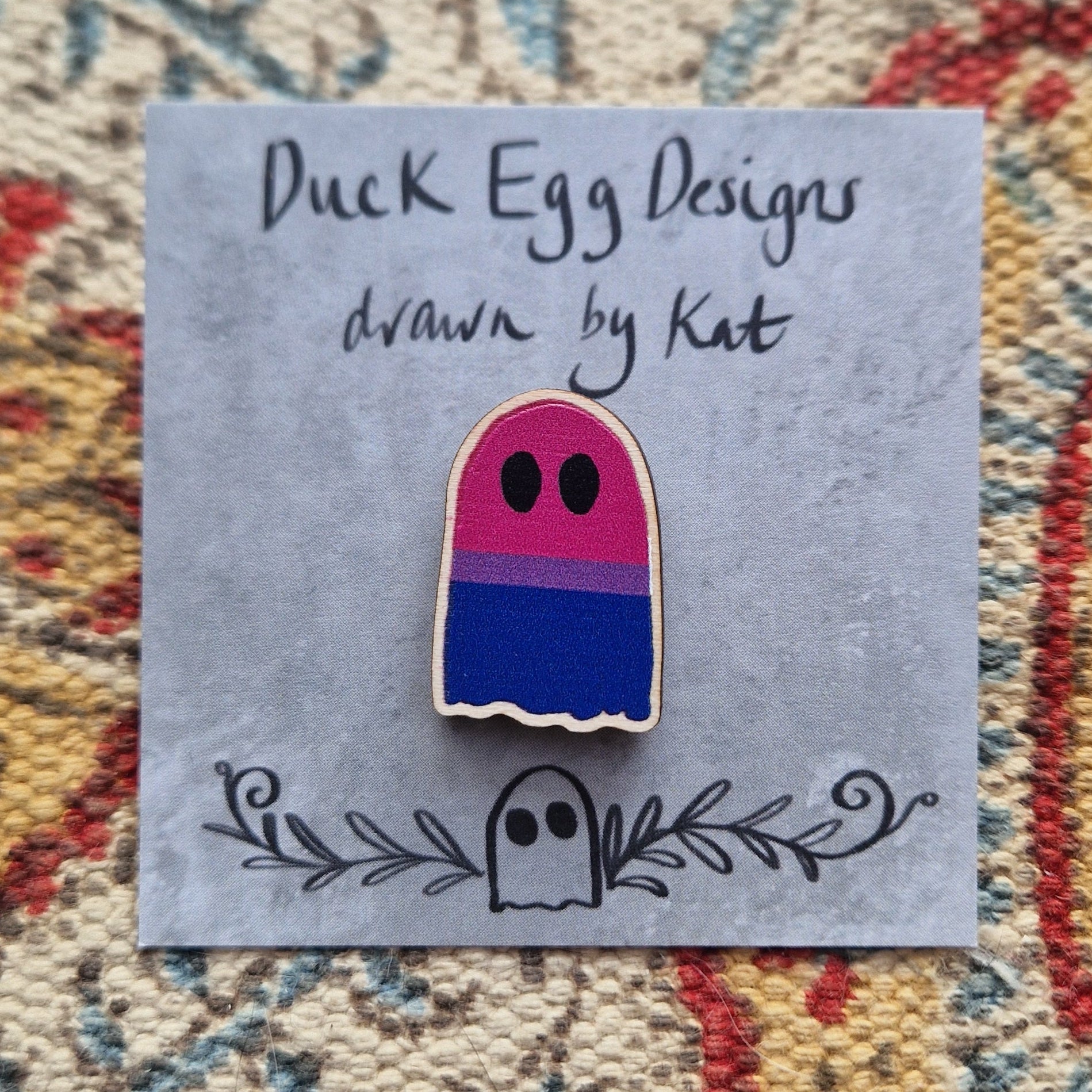 A ghost pin featuring the bisexual flag (with stripes of pink, purple and blue) sits on a grey backing card with the words 'Duck Egg Designs drawn by Kat' across the top. Behind the backing card is a patterned fabric background.