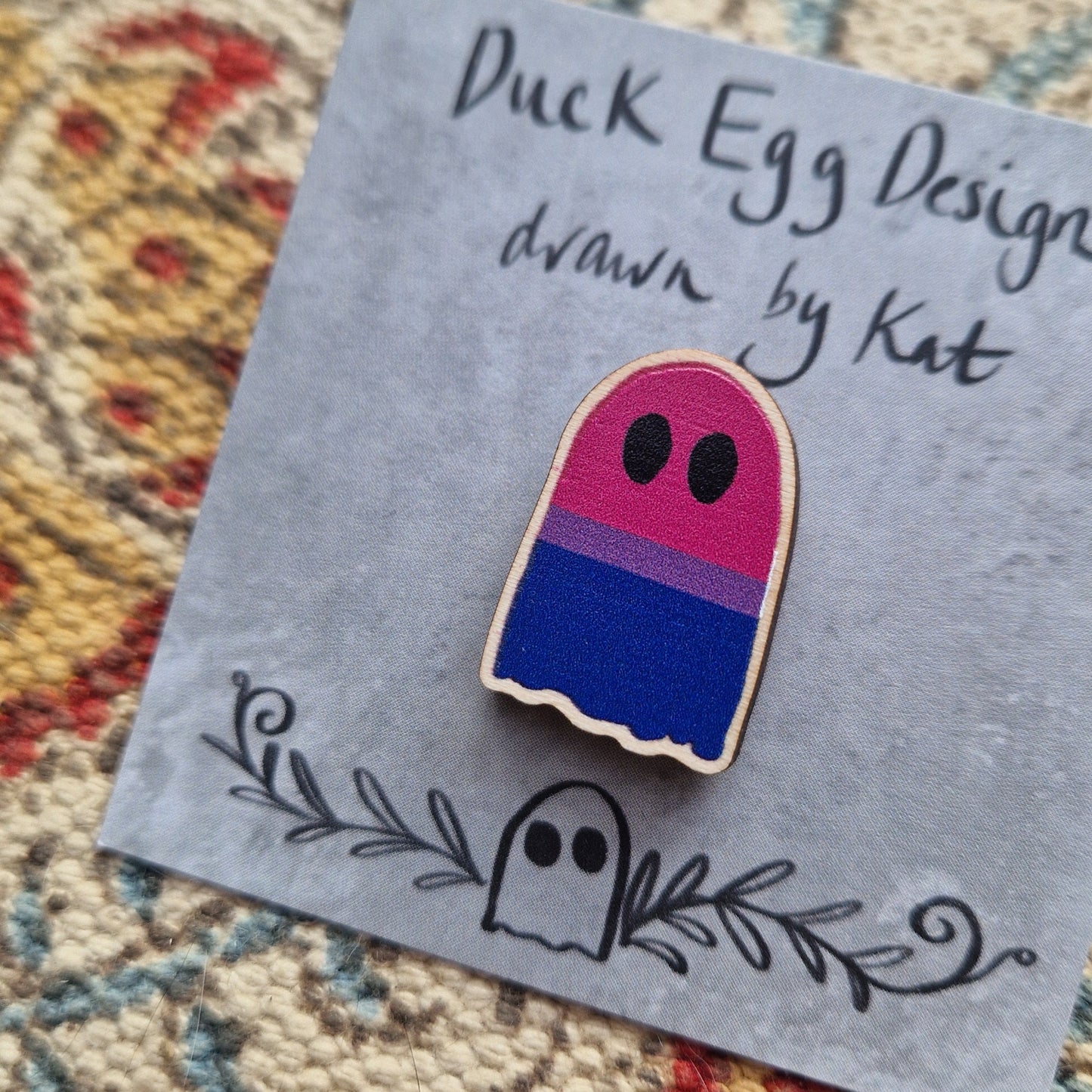 A ghost pin featuring the bisexual flag (with stripes of pink, purple and blue) sits on a grey backing card with the words 'Duck Egg Designs drawn by Kat' across the top. Behind the backing card is a patterned fabric background.