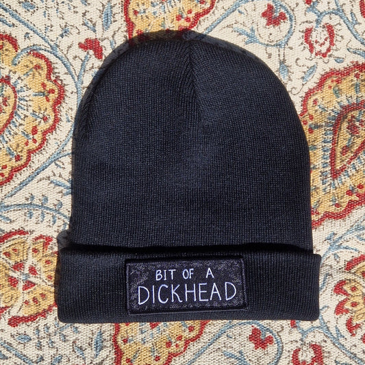A black beanie lays on a patterned fabric background. The beanie has a rectangular patch on it that reds 'Bit Of A Dickhead'.