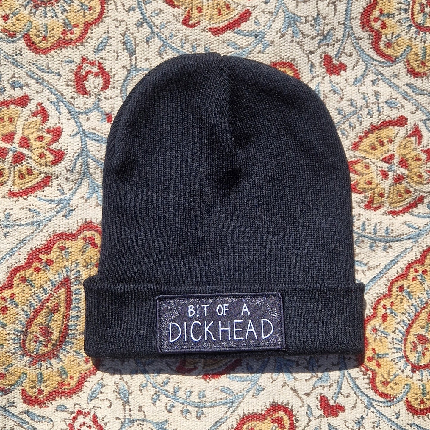 A black beanie lays on a patterned fabric background. The beanie has a rectangular patch on it that reds 'Bit Of A Dickhead'.