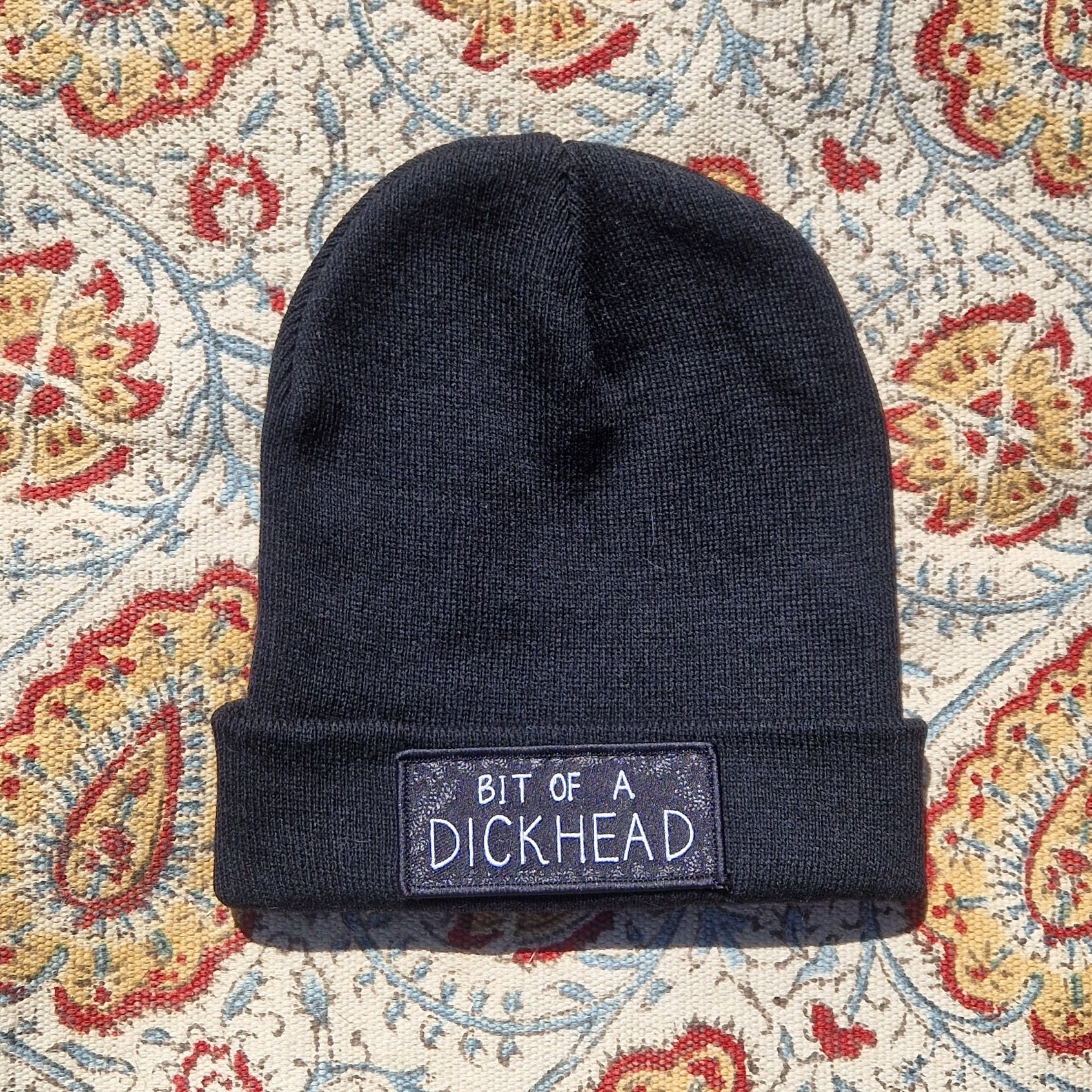 A black beanie lays on a patterned fabric background. The beanie has a rectangular patch on it that reds 'Bit Of A Dickhead'.