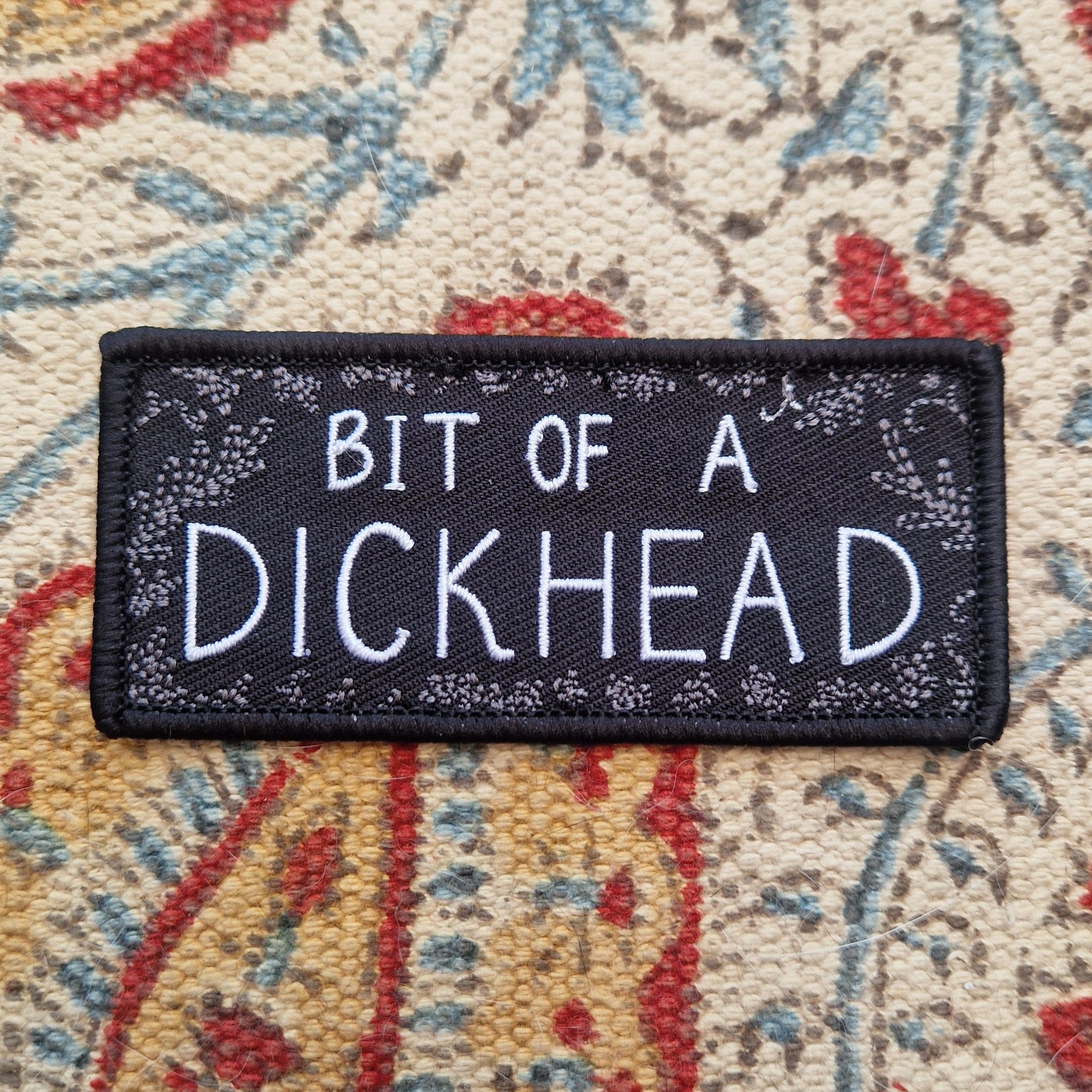 A black rectangular patch with the words 'BIT OF A DICKHEAD' across the middle in white text and a grey leafy frond design around the outside. The patch sits on a patterned fabric background.
