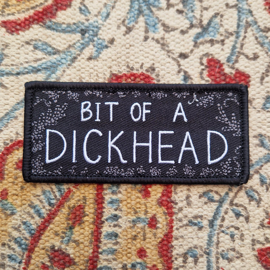 A black rectangular patch with the words 'BIT OF A DICKHEAD' across the middle in white text and a grey leafy frond design around the outside. The patch sits on a patterned fabric background.
