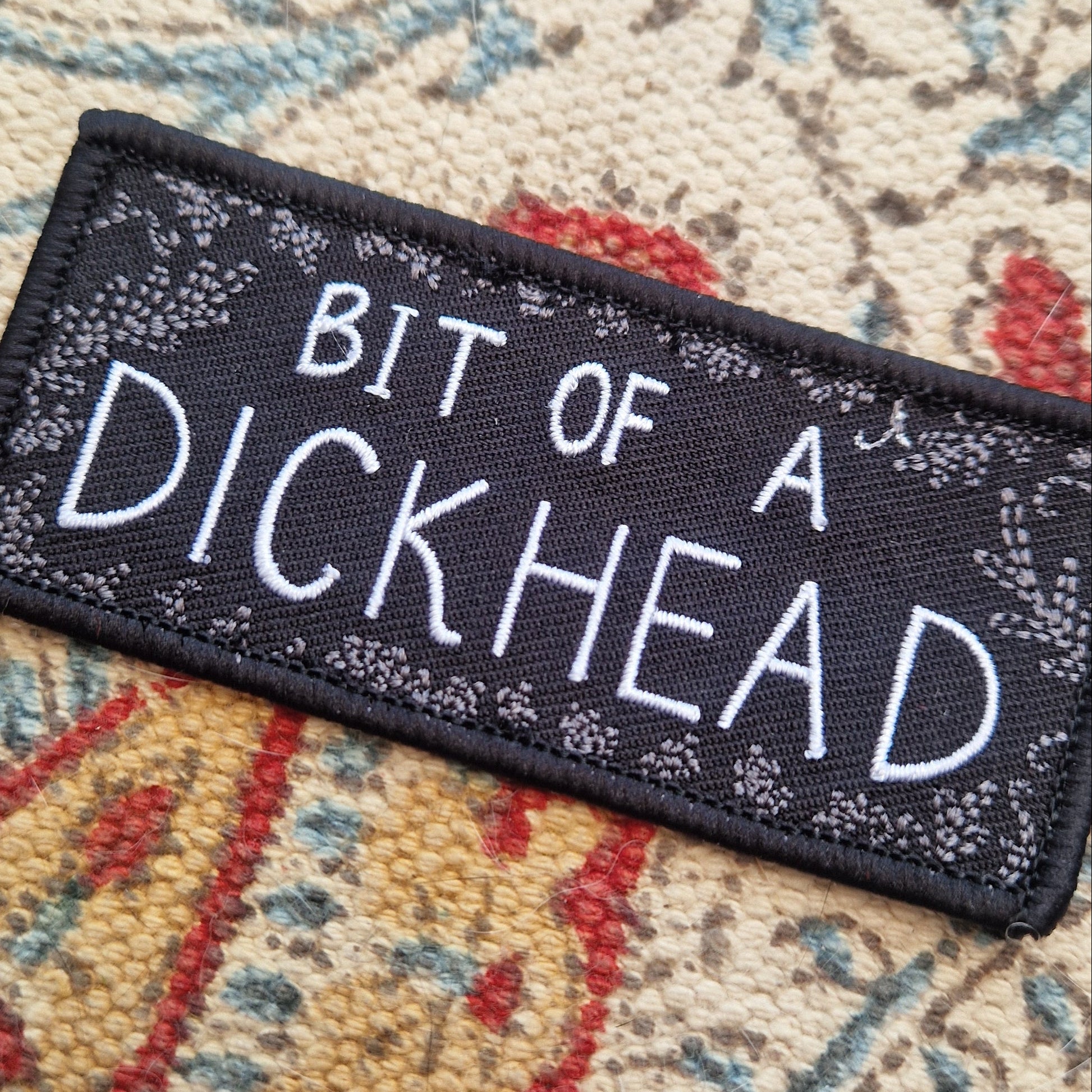 A close up of the black rectangular patch with the words 'BIT OF A DICKHEAD' across the middle in white text and a grey leafy frond design around the outside. The patch sits on a patterned fabric background.