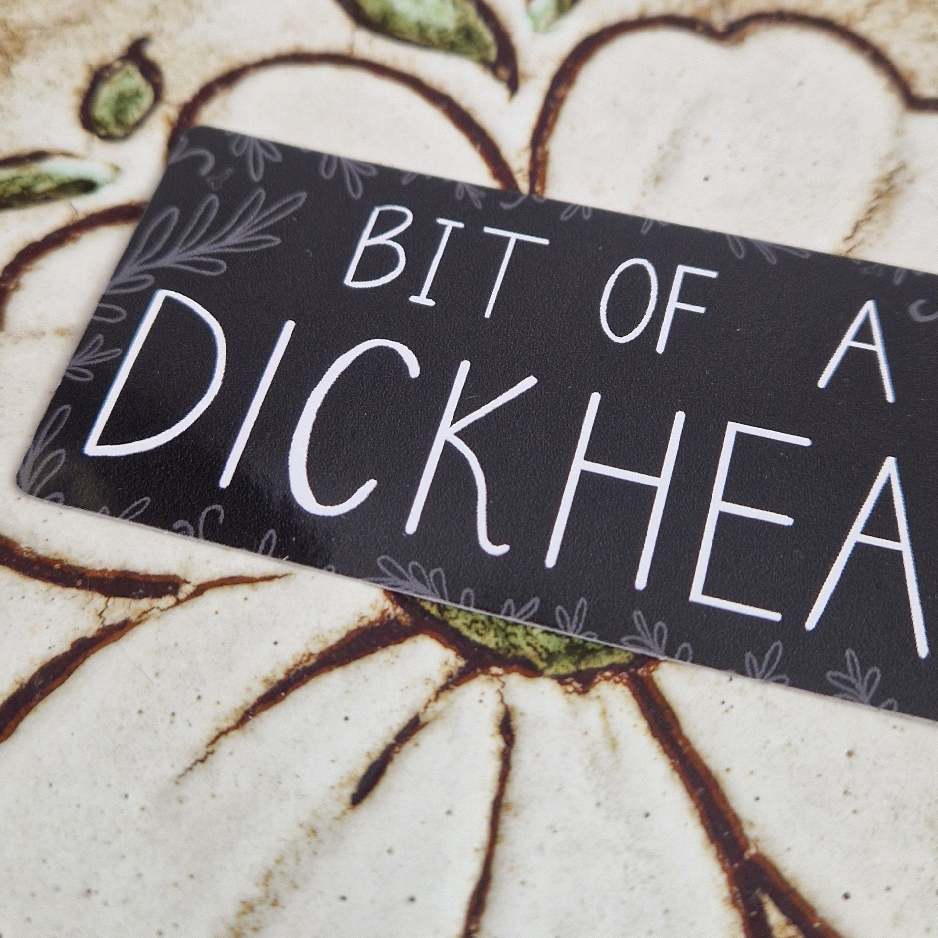 A closer look at the rectangular vinyl sticker with 'BIT OF A DICKHEAD' written in white handwriting with grey leafy tendrils growing in from the outside with some mini dick illustration dotted around too. The sticker sits on a floral tiled background.