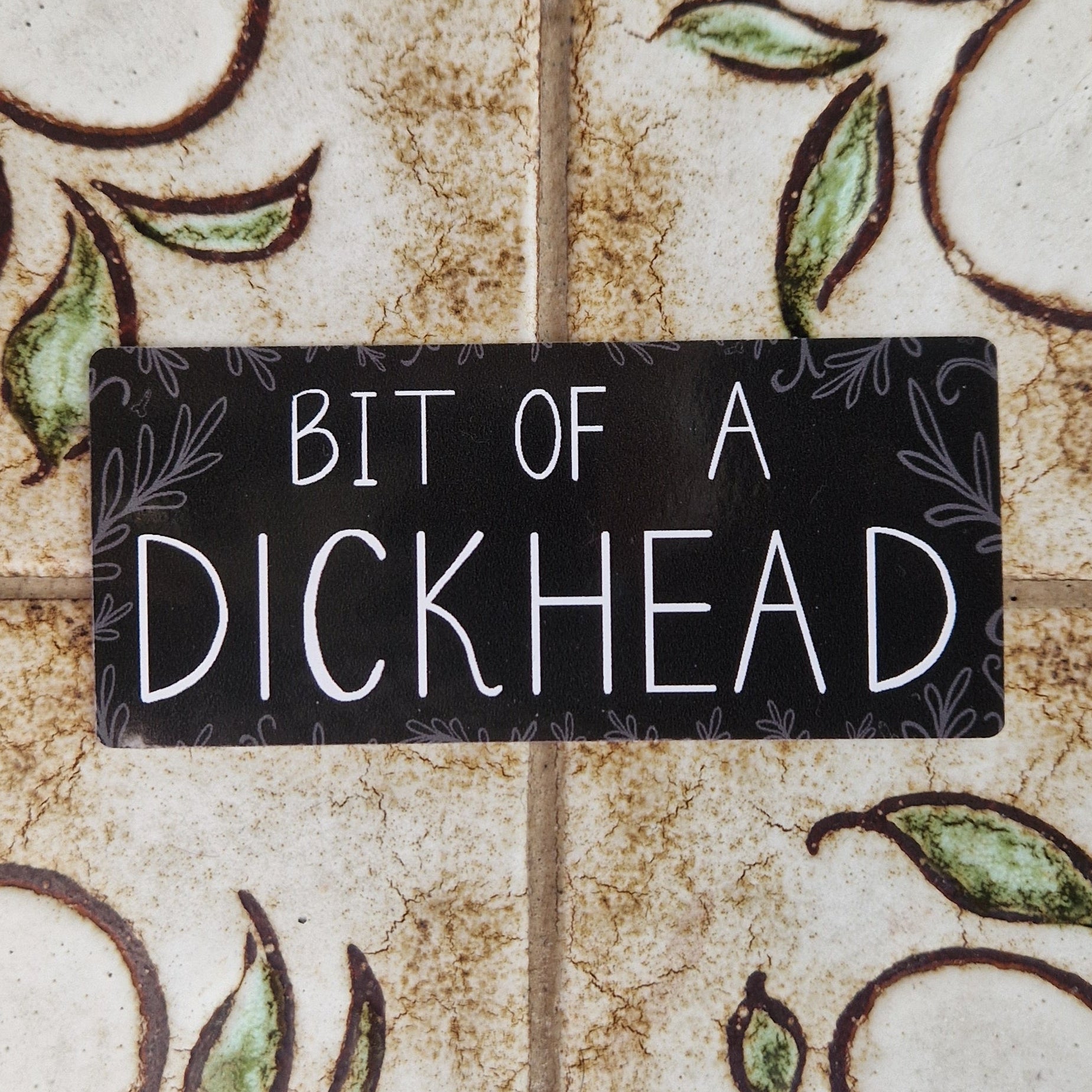 A black rectangular vinyl sticker on a floral tiled background, The sticker has the words 'BIT OF A DICKHEAD' in white handwriting surrounded by grey leafy fronds (and a few mini dick illustrations).