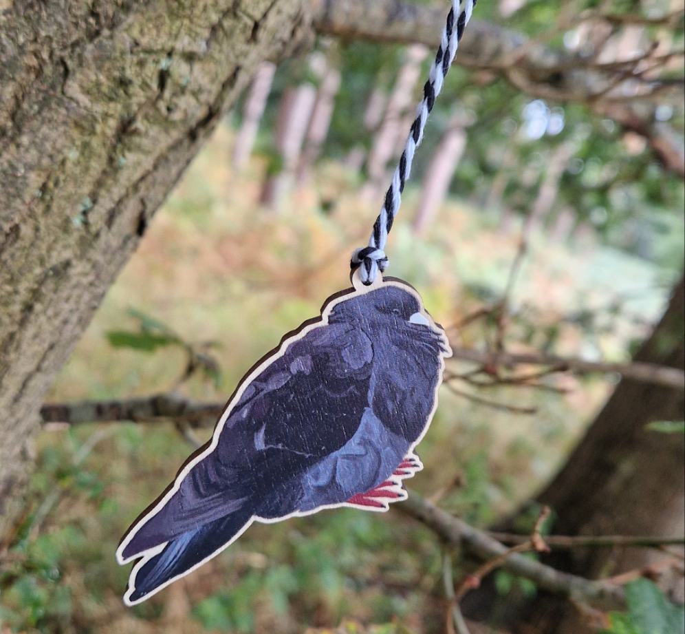 A black pigeon wooden decoration hangs from a white and black string in a green woods.