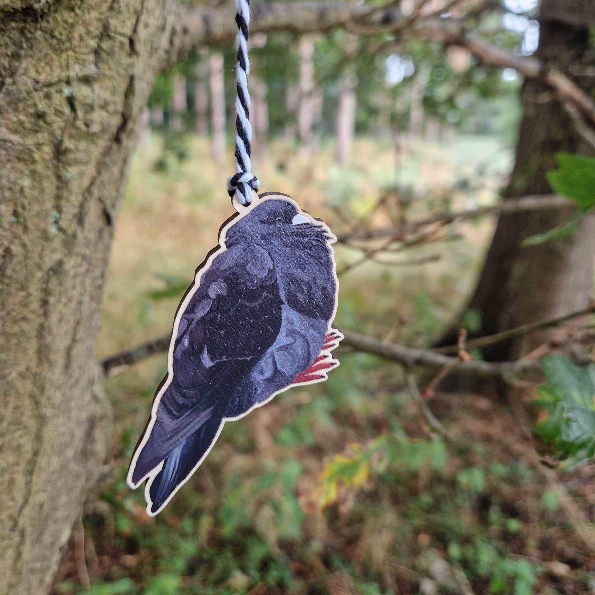 A black pigeon wooden decoration hangs from a white and black string in a green woods.