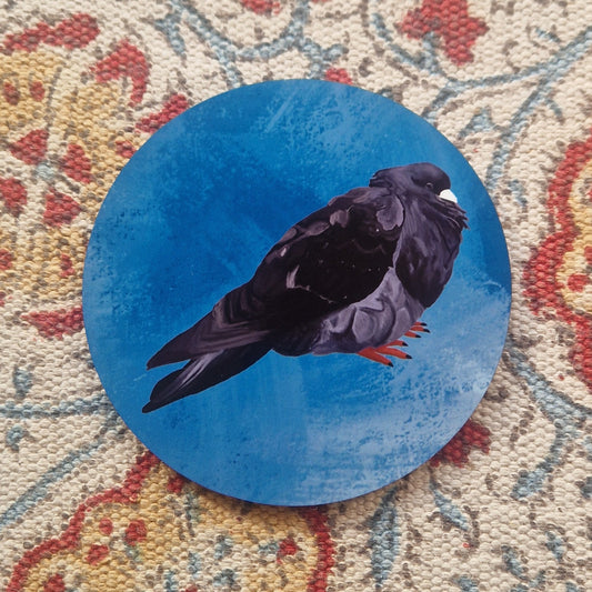 A blue coaster featuring a black round pigeon with a pale grey beak and orange feet sits on a patterned fabric background.