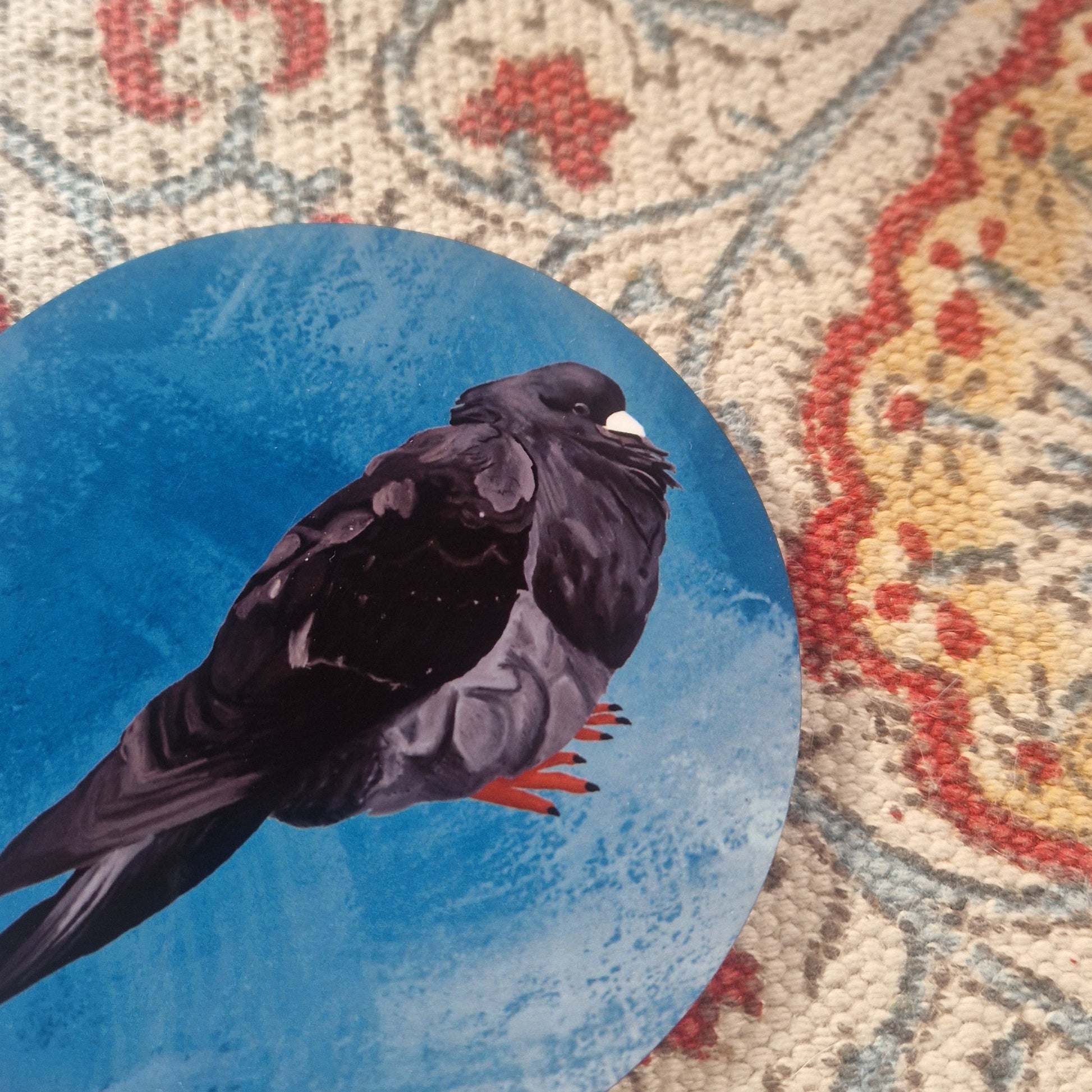 A close up of the blue coaster showing the detail in the lack pigeon illustration.Behind the coaster is a patterned fabric background.