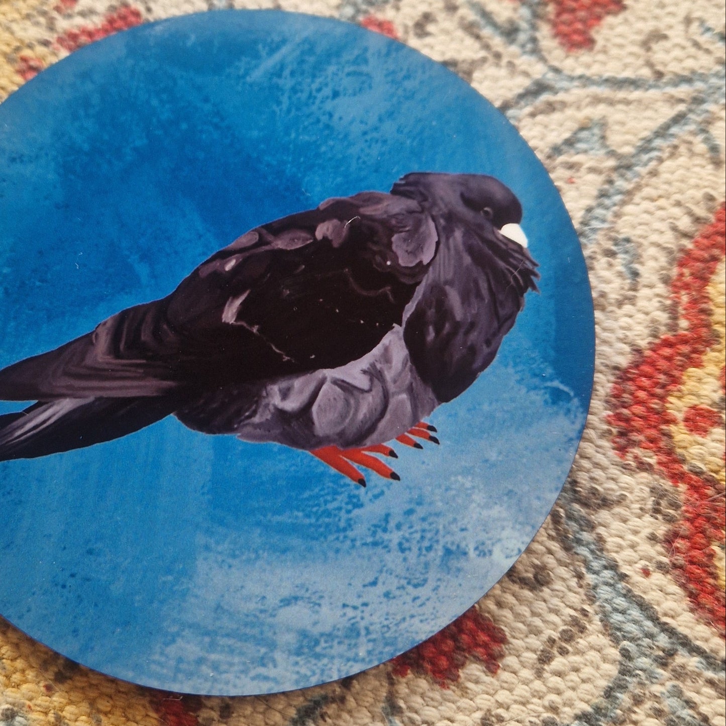 A close up of the round blue coaster showing the detail in the black fat pigeon illustration. Behind the coaster you can see a patterned fabric background.