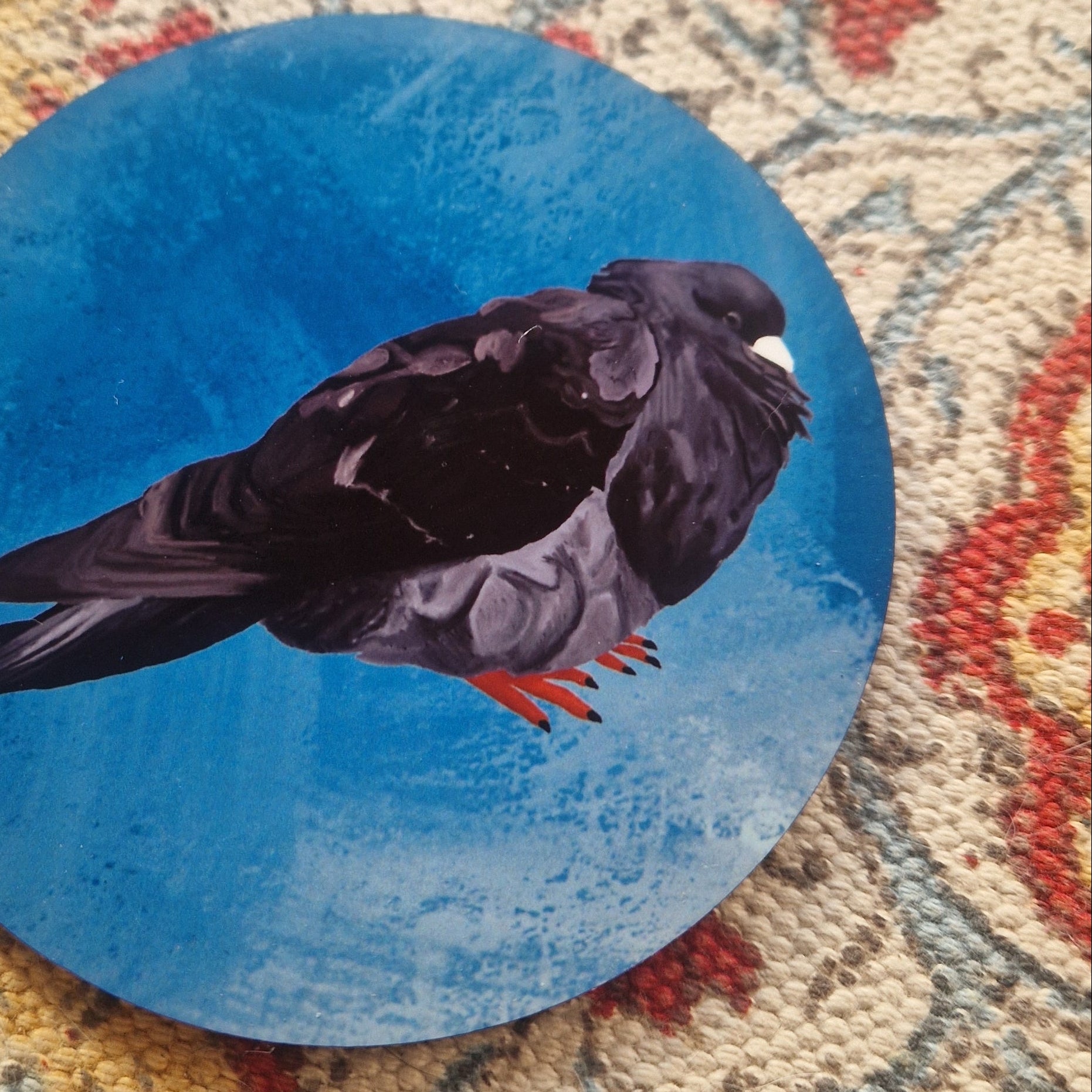 A close up of the round blue coaster showing the detail in the black fat pigeon illustration. Behind the coaster you can see a patterned fabric background.