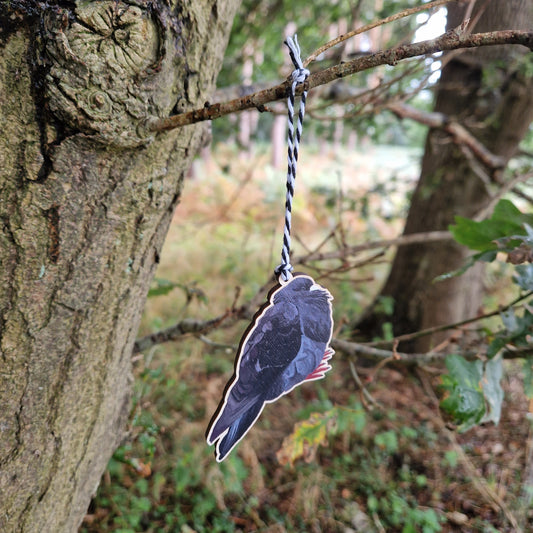A black pigeon wooden decoration hangs from a white and black string from a tree branch in a green woods.