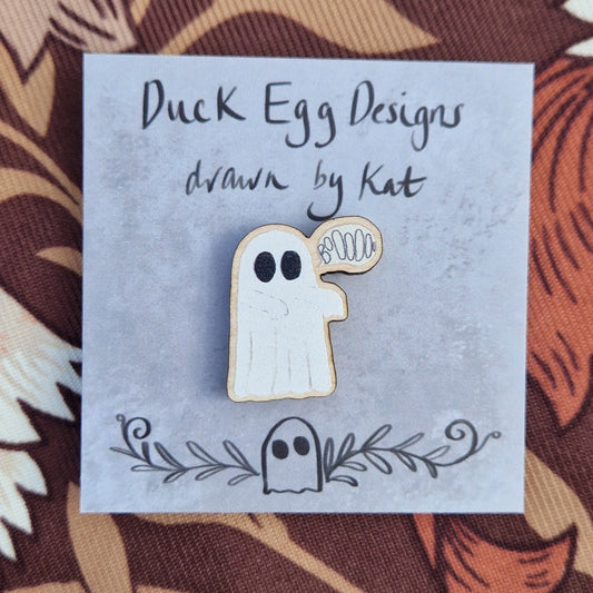 White ghost pin badge with an off white speech bubble with the word 'BoOOOOo' in it. The pin sits on a grey pin badge with the words 'Duck Egg Designs drawn by Kat' across the top. Behind the backing card is a patterned brown fabric.