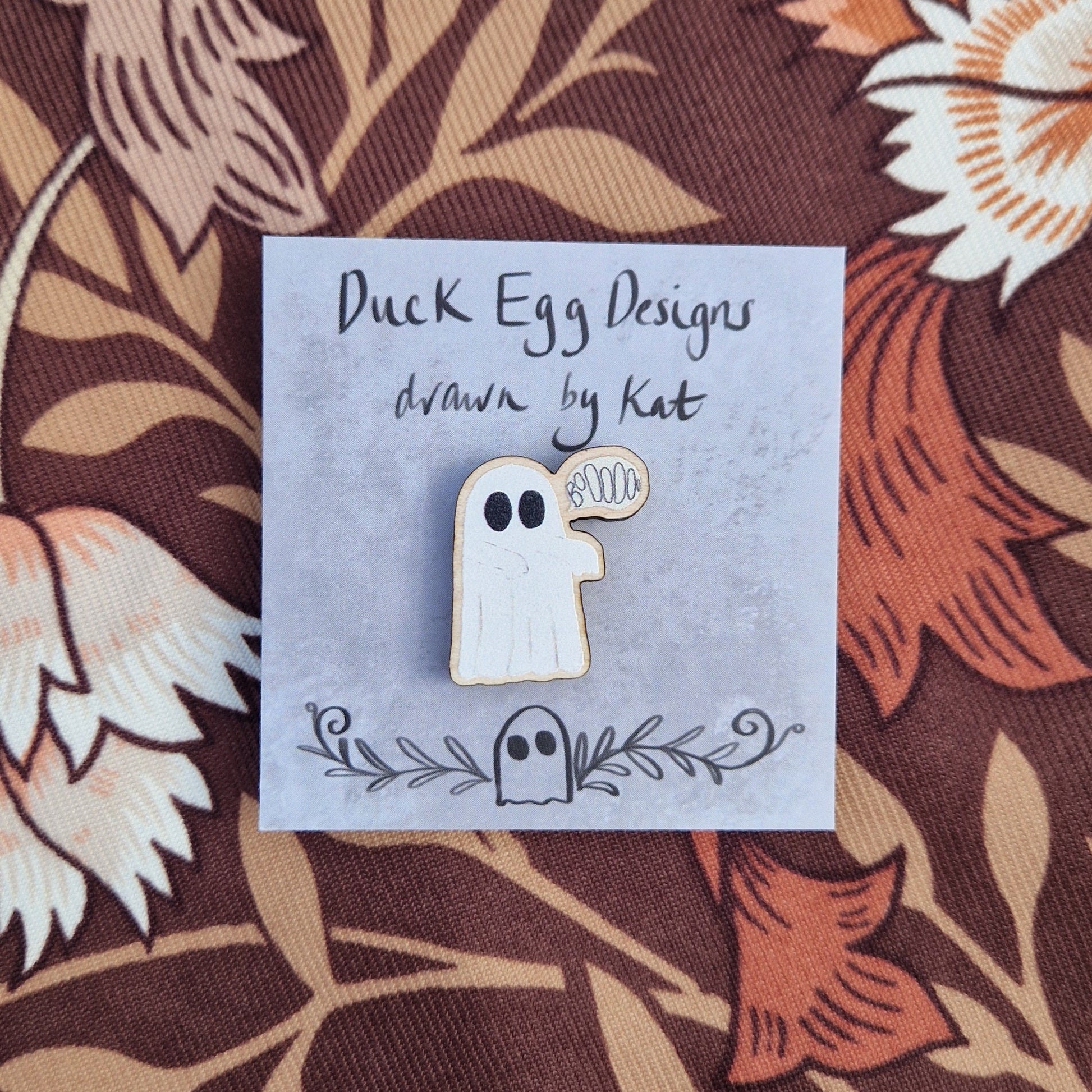 White ghost pin badge with an off white speech bubble with the word 'BoOOOOo' in it. The pin sits on a grey pin badge with the words 'Duck Egg Designs drawn by Kat' across the top. Behind the backing card is a patterned brown fabric.