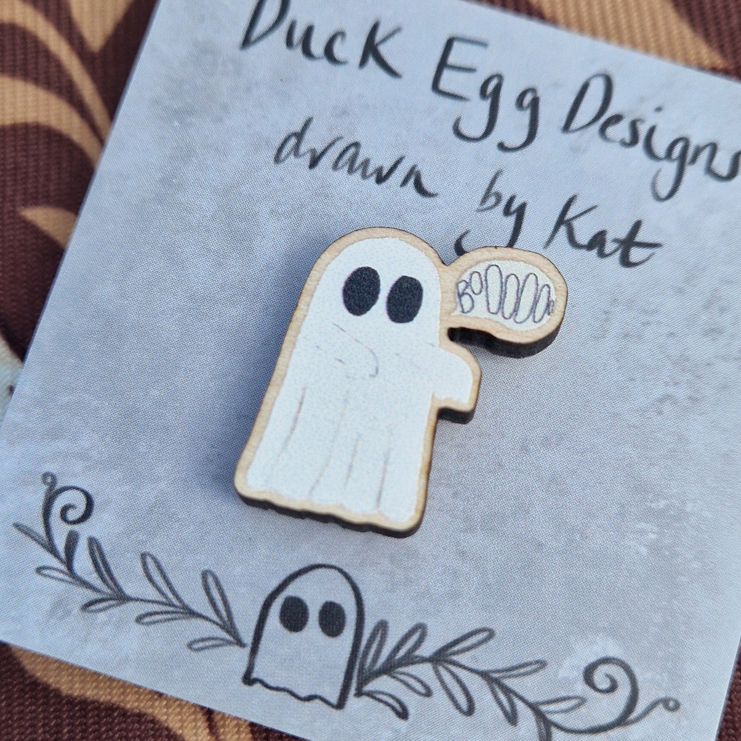 White ghost pin badge with an off white speech bubble with the word 'BoOOOOo' in it. The pin sits on a grey pin badge with the words 'Duck Egg Designs drawn by Kat' across the top. Behind the backing card is a patterned brown fabric.