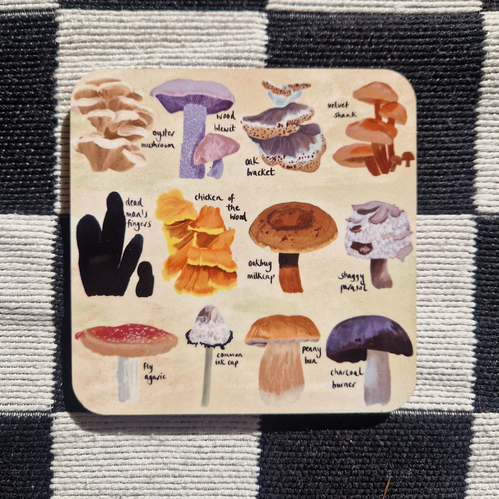 A square coaster featuring 12 different fungi illustrations on a cream background. Behind the coaster is a black and white checkered fabric background.